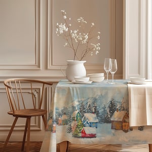 Könnte beinhalten: Eine rechteckige Tischdecke mit einem winterlichen Dorfmotiv, das verschneite Häuser, Tannenbäume und Berge zeigt. Der Tisch ist mit einem weißen Keramikkrug mit weißen Blumen, zwei weißen Schalen und zwei hohen Glasflöten gedeckt.