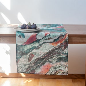 May include: A green, orange, and brown patterned table runner with a white plate and four figs on top of it. The runner is on a wooden table.