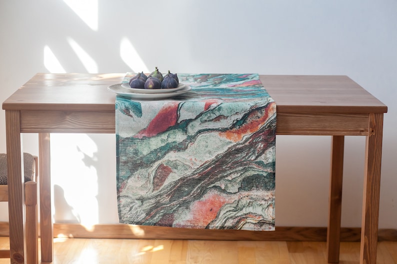 May include: A green, orange, and brown patterned table runner with a white plate and four figs on top of it. The runner is on a wooden table.