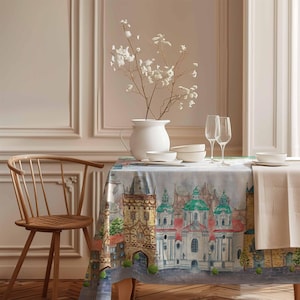 Könnte beinhalten: Eine weiße Tischdecke mit einem Aquarelldruck einer europäischen Stadtlandschaft. Die Tischdecke liegt auf einem Holztisch mit einem weißen Krug, zwei Weingläsern und weißen Schalen. Ein Holzstuhl mit einem geflochtenen Sitz befindet sich im Vordergrund.