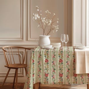 May include: A rectangular tablecloth featuring a vertical striped pattern with alternating bands of light green and a festive print of poinsettias, golden bells, pinecones, and red ribbons. The fabric drapes over a table set with white dishes.