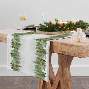 May include: A white table runner featuring a watercolour design of a snowy forest. The runner is decorated with green pine trees and white snowflakes. The runner is placed on a wooden table with a festive centrepiece of greenery, candles, and fairy lights.