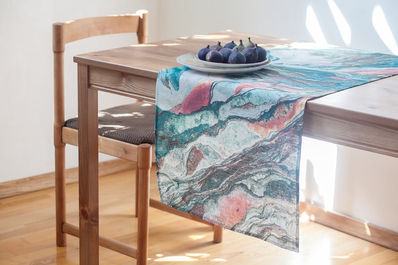 May include: A teal, pink, and brown abstract patterned table runner on a wooden table with a plate of figs.