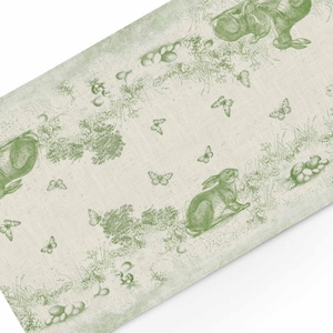 Puede incluir: Una tela rectangular blanquecina con un estampado verde de toile de Jouy. El diseño presenta conejos, mariposas y follaje. La tela es probablemente un camino de mesa o un paño decorativo.