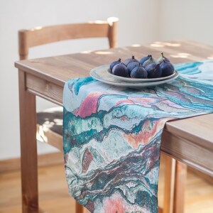 May include: A teal, pink, and brown marble-patterned table runner on a wooden table with a plate of figs.