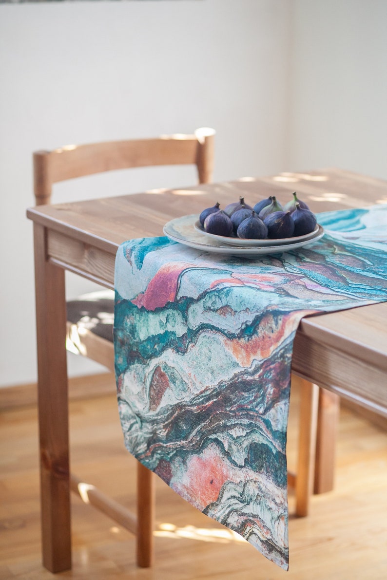 May include: A teal, pink, and brown marble-patterned table runner on a wooden table with a plate of figs.