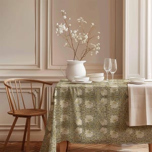 Pode incluir: Uma toalha de mesa floral verde e branca com um padrão delicado. A toalha de mesa está posta sobre uma mesa de madeira com um jarro branco, tigelas e copos. Uma cadeira de madeira com um assento de vime está em primeiro plano.
