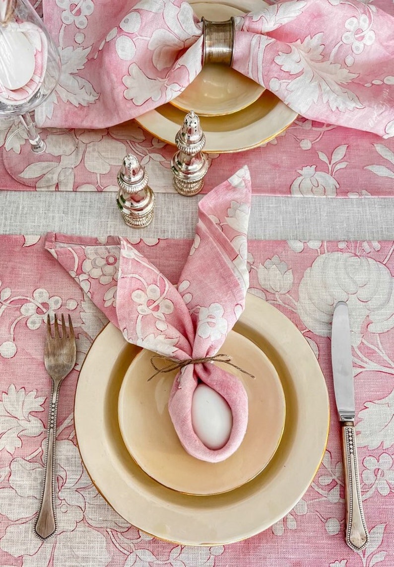 May include: A pink floral tablecloth with a place setting featuring a white egg wrapped in a pink napkin, a white plate, a gold-rimmed plate, a silver fork, and a silver knife.