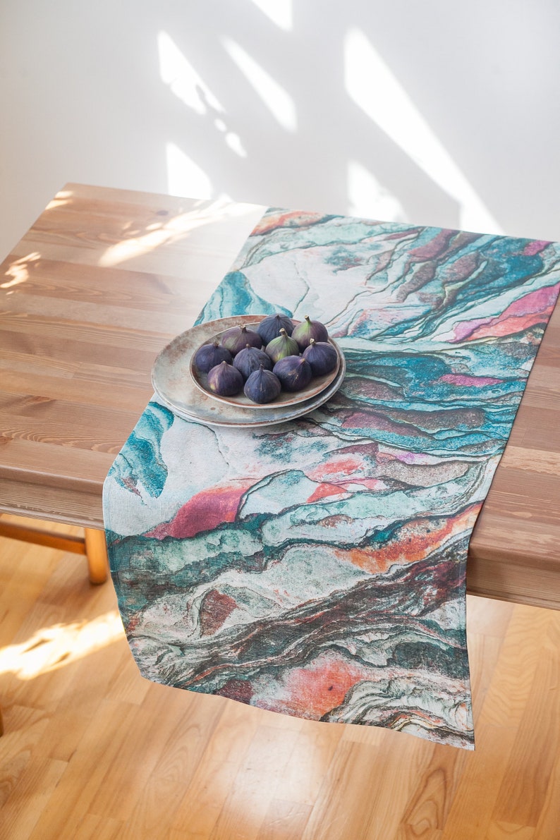 May include: A colorful table runner with a marbled pattern in shades of blue, green, pink, and orange. A plate of dark purple figs sits on top of the runner.