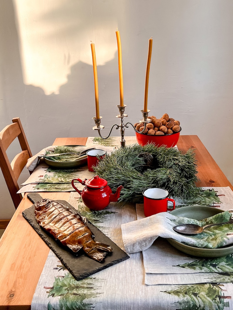 May include: A table setting with a smoked fish on a black slate platter, a red teapot, two red mugs, a green bowl, a green wreath, and a bowl of walnuts. The table is covered in a linen tablecloth with a green and white pattern.