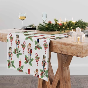 May include: White table runner with a repeating pattern of nutcracker soldiers, candy canes, holly, and candles. The runner is on a wooden table with a centerpiece of greenery and candles.