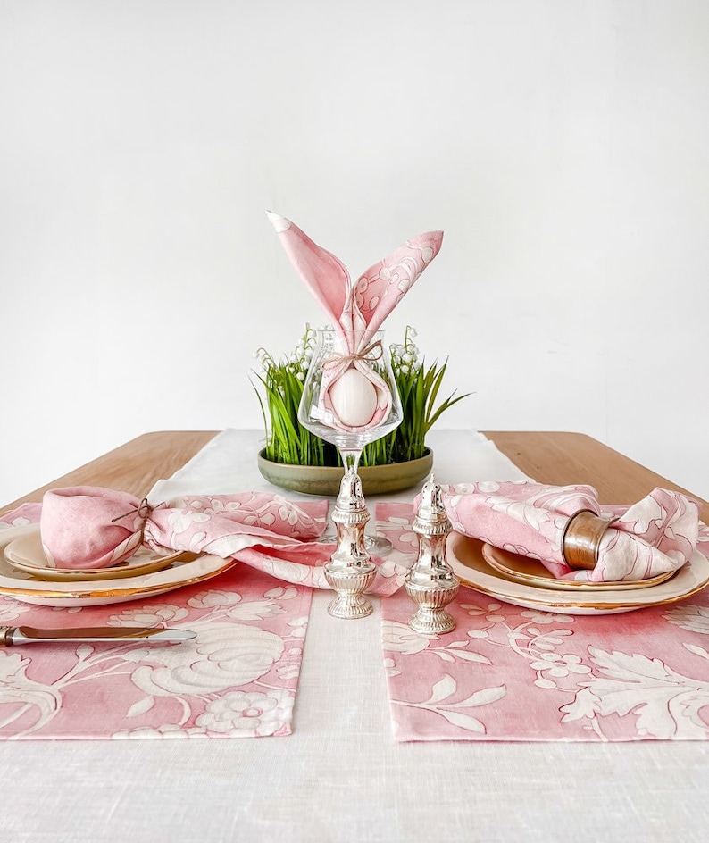 May include: Pink floral table runner with two place settings. Each place setting includes a pink floral placemat, a white plate, a pink napkin folded into a bunny shape, and a silver salt and pepper shaker. A glass vase with a white egg and green grass is in the centre of the table.