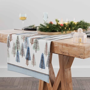 Puede incluir: Un camino de mesa blanco con un borde azul y un patrón de árboles de estilo acuarela en tonos azules, verdes y marrones. El camino de mesa está sobre una mesa de madera con un centro de mesa de vegetación y velas.