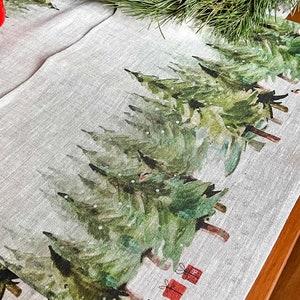 May include: A linen table runner with a watercolor design of green pine trees and red gift boxes. The runner is on a wooden table.
