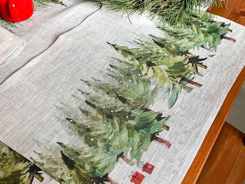 May include: A linen table runner with a watercolor design of green pine trees and red gift boxes. The runner is on a wooden table.