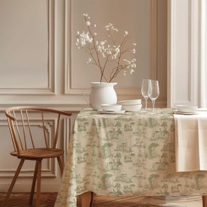 May include: A rectangular tablecloth featuring a repeating green toile de jouy pattern of horses and trees on a cream background. The fabric drapes over a table set with a white pitcher containing white flowers, two wine glasses, and white bowls.