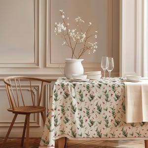 Könnte beinhalten: Eine rechteckige Tischdecke mit einem sich wiederholenden Muster aus grünen Blättern und kleinen roten Beeren auf cremefarbenem Grund, drapiert über einen Tisch mit weißem Krug, weißen Schalen und zwei klaren Weingläsern.
