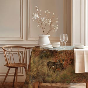 May include: A rectangular tablecloth featuring a detailed forest scene with two deer standing among autumn foliage in shades of brown, orange, and green. The table is set with a white ceramic pitcher containing white flowers, bowls, and two wine glasses.