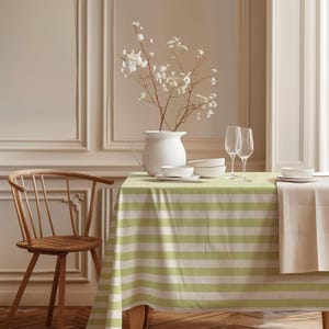 May include: A dining table set with a green and white striped tablecloth, white dishes, and wine glasses. A white vase holds delicate white flowers. A wooden chair is visible on the left.