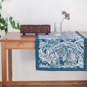 May include: A blue and white floral table runner with a white background and a blue floral pattern. The runner is on a wooden table with a wooden box and a glass vase with dried flowers.