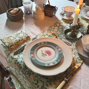 Könnte beinhalten: Ein gedeckter Tisch mit einer Tischdecke mit Blumenmuster, mit Tellern, Besteck und einem Brotkorb. Ein kleiner Teller mit einem rosa Blumendesign liegt auf einem größeren weißen Teller. Angezündete Kerzen und Teetassen tragen zum Ambiente bei.