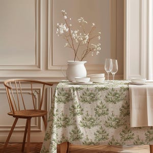 May include: A dining table set with a tablecloth featuring a green tree pattern. White dishes, wine glasses, and a white vase with white flowers are on the table. A wooden chair is on the left. The table is set for a meal.