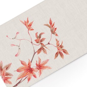 May include: A rectangular table runner made of light beige fabric featuring a botanical illustration of a Japanese maple branch. The branch displays vibrant red and orange palmate leaves along with several light pink winged maple seeds.