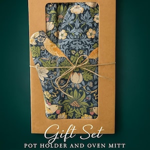 Può includere: Un set regalo con un guanto da forno e un sottopentola a fantasia floreale, presentato in una scatola di cartone marrone legata con spago. Il tessuto ha uno sfondo blu scuro con uccelli gialli e fiori bianchi. Il testo "Gift Set" è visibile.