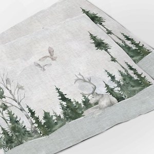 以下が含まれることがあります： 鹿、フクロウ、松の木が描かれた雪降る森の風景の水彩画。イラストは薄いグレーのリネン生地に印刷されています。