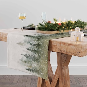 May include: A white table runner with a watercolor design of a snowy forest. The runner is placed on a wooden table with a festive centerpiece of greenery, candles, and lights.