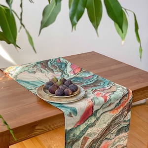 May include: A colorful table runner with a marbleized pattern in shades of green, brown, pink, and white. The runner is laid on a wooden table and features a plate of fresh figs.