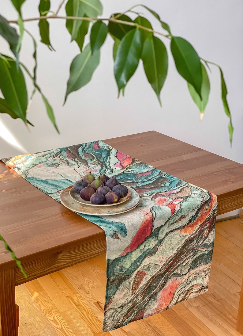 May include: A colorful table runner with a marbleized pattern in shades of green, brown, pink, and white. The runner is laid on a wooden table and features a plate of fresh figs.