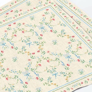 Peut inclure: Un set de table rectangulaire en tissu sur fond beige, orné d'un motif répétitif de petites fleurs roses, jaunes et bleues, de feuilles vertes et de rubans bleu clair. La bordure se compose d'une fine ligne bleue et d'un motif de vigne verte.