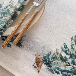 May include: A white linen placemat with a watercolor design of a fox in a snowy forest. The placemat has a green and blue color scheme and features a brown and silver fork and knife.