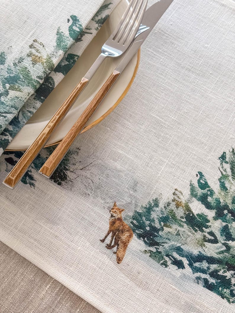 May include: A white linen placemat with a watercolor design of a fox in a snowy forest. The placemat has a green and blue color scheme and features a brown and silver fork and knife.