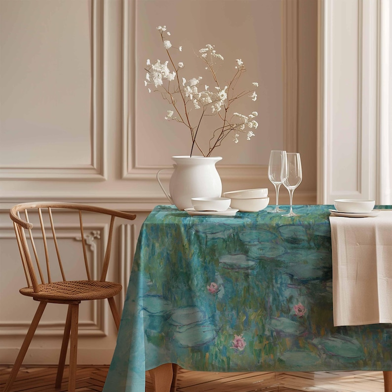 Op de afbeelding: Een blauw en groen tafelkleed met een waterleliemotief. Het tafelkleed ligt op een houten tafel met een witte kan, kommen en glazen. Een houten stoel staat op de voorgrond.
