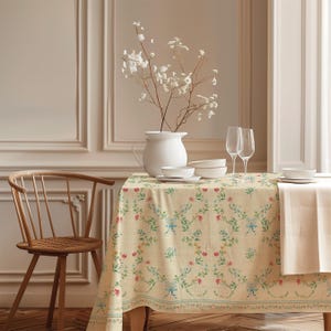 Può includere: Una tovaglia rettangolare con base color crema e un motivo ripetuto di fiori rosa e gialli, foglie verdi e fiocchi azzurri. Il tessuto copre un tavolo apparecchiato con una brocca bianca, ciotole bianche e due bicchieri alti.