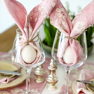 May include: Two pink floral patterned napkins are tied around white eggs in clear wine glasses. The glasses are on a pink and white floral tablecloth.
