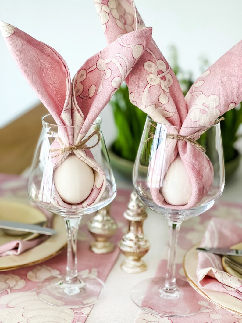 May include: Two pink floral patterned napkins are tied around white eggs in clear wine glasses. The glasses are on a pink and white floral tablecloth.