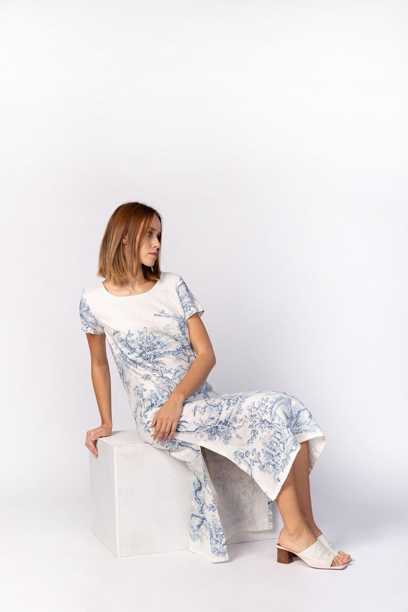 May include: A woman wearing a white dress with a blue floral pattern sits on a white cube. She is wearing white heeled sandals.