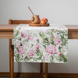 Könnte beinhalten: Ein weißer Tischläufer aus Leinen mit einem floralen Muster aus rosa Pfingstrosen und grünen Blättern. Der Läufer liegt auf einem Holztisch mit einer braunen Keramiktasse und einem Pfirsich.