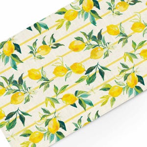May include: A white linen table runner with a yellow and green lemon print. The lemons are arranged in a repeating pattern on a background of yellow stripes.