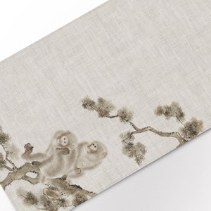 May include: A beige linen placemat featuring a delicate black and white illustration of two monkeys sitting on a branch of a pine tree.