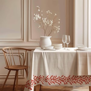 Könnte beinhalten: Eine weiße Tischdecke mit einem roten Beerenrand, gedeckt für ein Abendessen mit zwei Weingläsern, Tellern und einer Vase mit weißen Blumen.