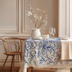 Könnte beinhalten: Eine rechteckige Tischdecke mit einem blau-weißen floralen Rankenmuster, die über einen Tisch drapiert ist. Das Design besteht aus komplexen, fließenden blauen botanischen Motiven, die auf einen cremefarbenen Stoff gedruckt sind.