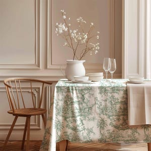 May include: A white tablecloth with a green toile pattern featuring a repeating design of trees, houses, and figures. The tablecloth is laid on a wooden table with a wooden chair and a white vase with dried flowers.