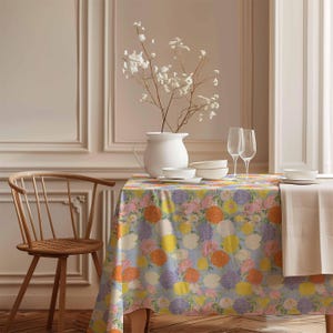 May include: A floral tablecloth with a light blue background and a variety of colourful flowers, including orange, yellow, pink, and purple. The tablecloth is laid on a wooden table with a white jug and two wine glasses.