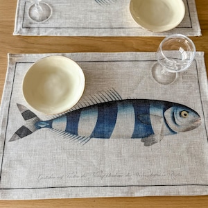 Peut inclure: Un set de table en lin avec un motif de poisson rayé bleu et blanc. Le set de table a une bordure noire et le texte "Geschmack auf  Tisch der  Fischhandlung  die  Meeresschätze  in  Berlin" est imprimé en bas.