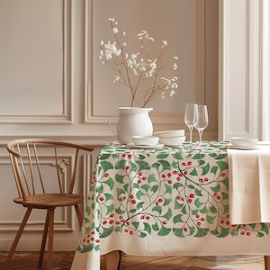 Op de afbeelding: Een rechthoekig tafelkleed met een herhalend patroon van groene ginkgobladeren en kleine rode bessen op een crèmekleurige achtergrond. Het tafelkleed ligt over een tafel gedekt met een witte kan, witte kommen en twee heldere glazen.