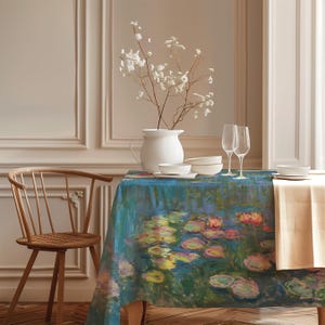 Op de afbeelding: Een eettafel met een blauw tafelkleed met een waterlelie schilderij. De tafel is gedekt met wit servies, wijnglazen en een witte vaas met witte bloemen. Links staat een houten stoel.